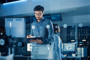 In Technology Research Facility: Chief Engineer Stands in the Middle of the Lab and Uses Tablet Computer. Team of Industrial Engineers, Developers Work on Engine Design Use Digital Whiteboard and Computers