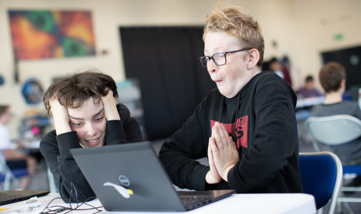 Two children sat at a desk with a laptop with excited expressions. 