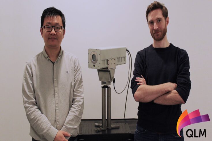 Xaio Ai, chief technical officer and Alex Dunning, head of engineering at QLM stand either side of their gas imaging camera. They are looking at the camera and smiling.