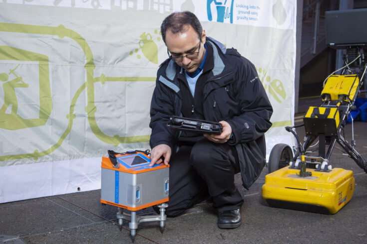 Dr Farzad Hayati kneels next to a current version of ground penetrating radar (GPR) at the National Quantum Technology Hub at Birmingham University. The GPR is a small silver box with orange bands at the top and bottom of the box.