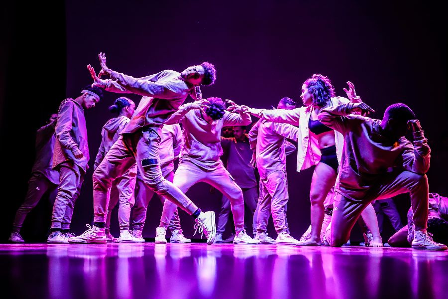 Thirteen hip-hop dancers on stage, lit up in a purple light.