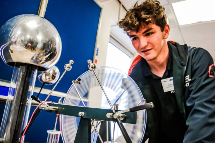 Apprentice wearing a dark blue coat and dark blue t-shirt with bright orange outlines inspecting a van de graff generator