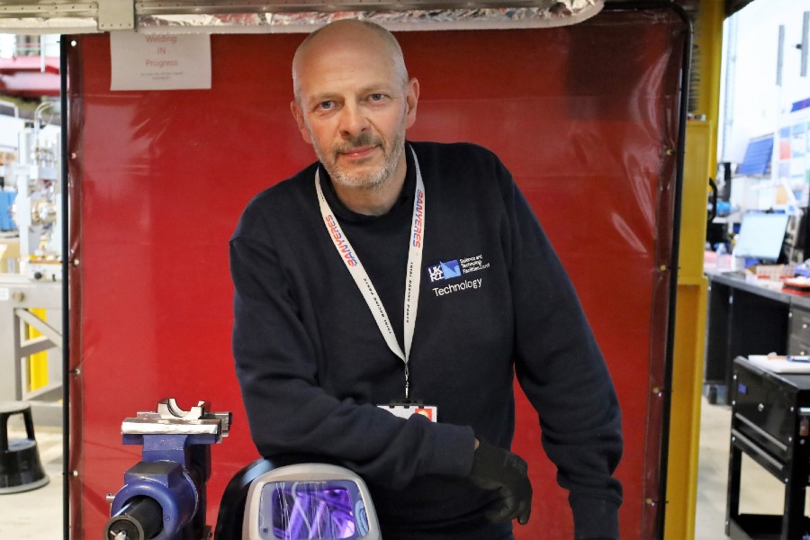 STFC welding expert John Hassall smiles at the camera. He is wearing an STFC branded blue sweater and leaning on his welding mask. In the background you can just see part of the Engineering Technology Centre at STFC Daresbury Laboratory. 