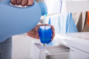 A close-up shot of a person filling the lid of a blue detergent bottle with detergent liquid next to a washing machine. Clothes hanging on a rack behind it.