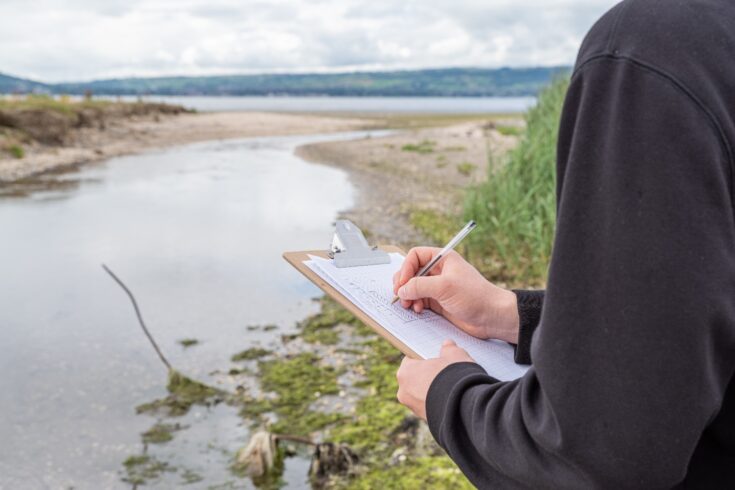 A researcher making field observations of a river and coastline with a clipboard and pen.