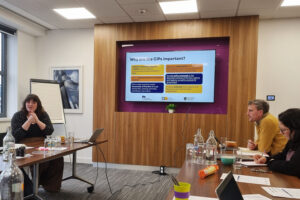 People in a meeting room. One person is leading discussion while the others listen.