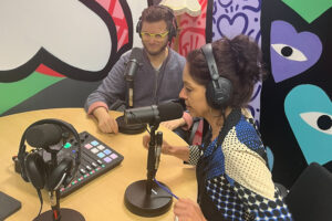 Georgina and Gaston, who are sitting at a table wearing headphones and speaking into large podcast microphones. 