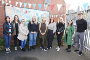 A group of people, who are standing outside a building that is decorated with a large mosaic and hanging bunting. 