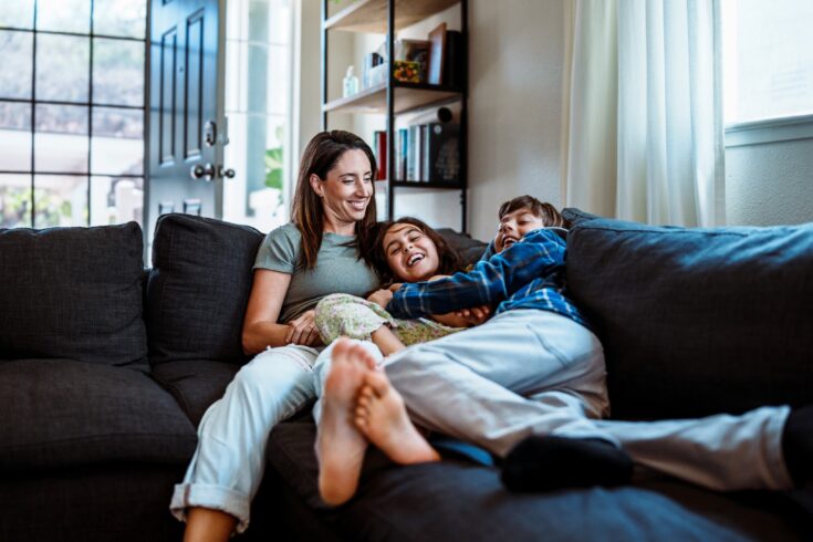 A mother and two playful children laughing together on a sofa.