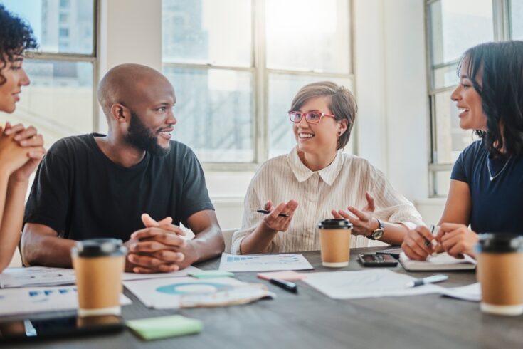 A group of happy colleagues sitting at a table and discussing work with coffees and papers.