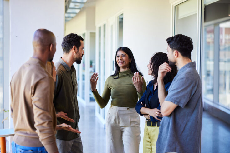 Young businesswoman talking and laughing with a group of diverse coworkers while standing together in the corridor of an office.