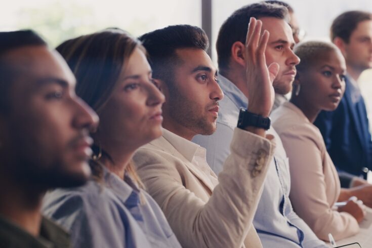 A group of professionals in a meeting, one man with his hand raised to make a comment.