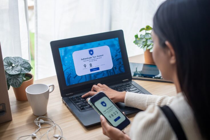 A young woman at a desk using two-factor authentication on her phone and laptop.