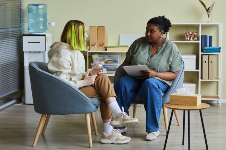 Female social worker talking to a teenage girl while they are both seated on armchairs in an office.