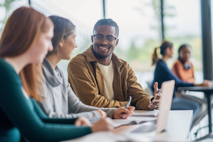 A small group of university students are seen sitting side-by-side at a desk in class as they work away on an assignment. They each have books open in front of them as they collaborate and share ideas.