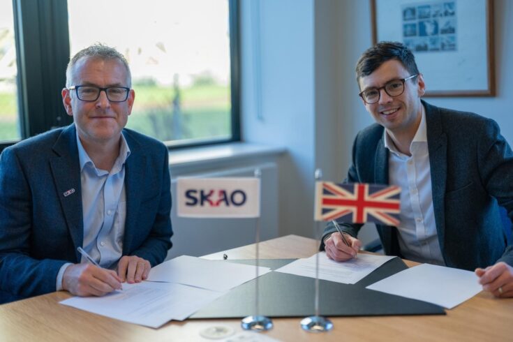 Two men sitting at a desk, smiling and signing papers, with a small Union Jack flag and a flag showing the SKAO logo on the desk in the foreground.