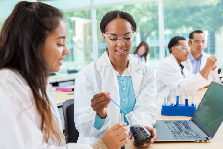 Confident diverse female scientists work on research together. They are dropping liquid into a beaker and then recording the findings on a laptop computer. Scientists are working in the background.