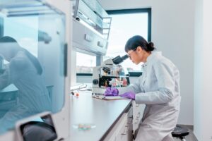 A researcher wearing a white lab coat looking into a microscope and writing notes on a note pad in a lab