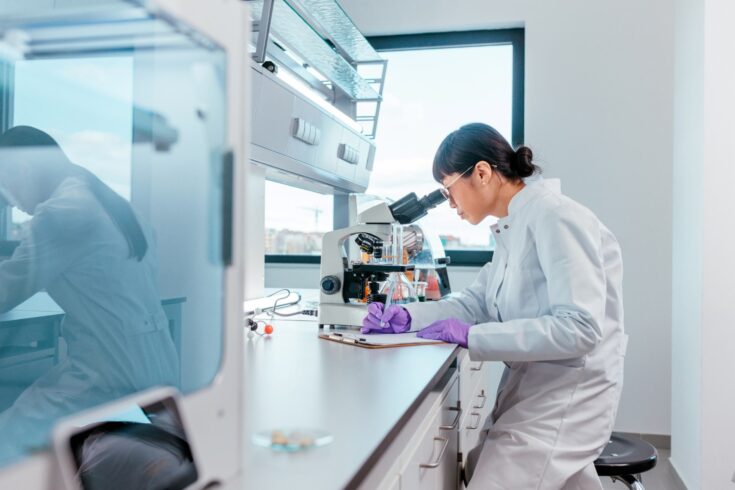 A researcher wearing a white lab coat looking into a microscope and writing notes on a note pad in a lab