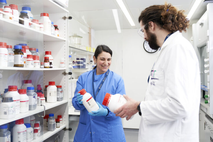 Two scientists in a lab holding lab storage bottles and talking with each other