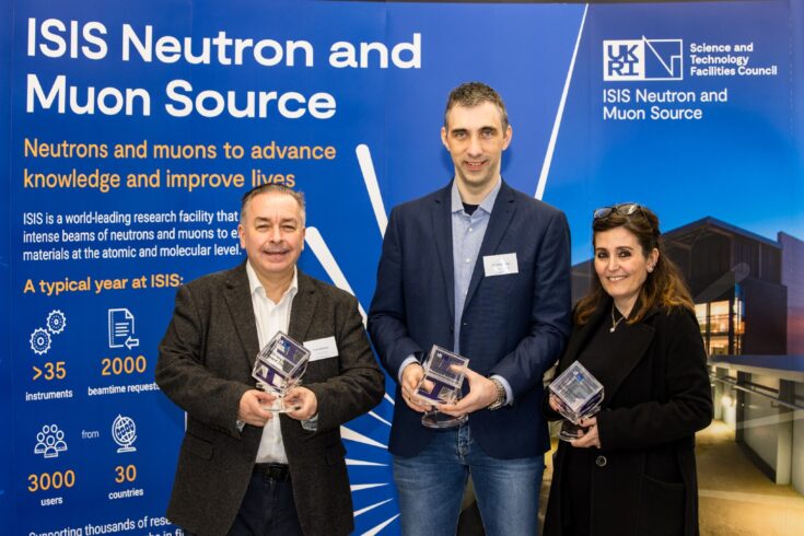 Three impact award winners holding their award trophies. They are stood in front of an ISIS Neutron and Muon Source banner about the world-leading facility using neutrons and muons to advance knowledge and improve lives