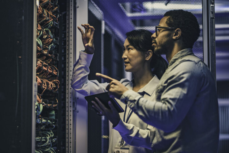 Side view of a female Asian IT engineer explaining network connections in a server to a novice African-American technician. ‎