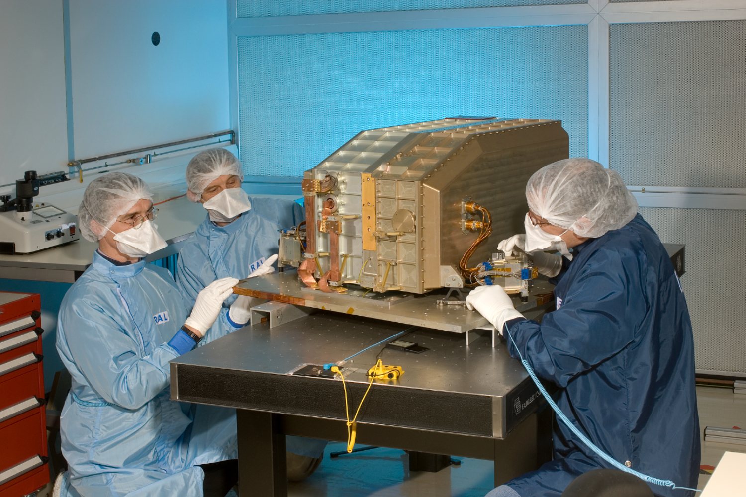 Three researchers wearing masks and gloves sitting around an instrument in a lab.